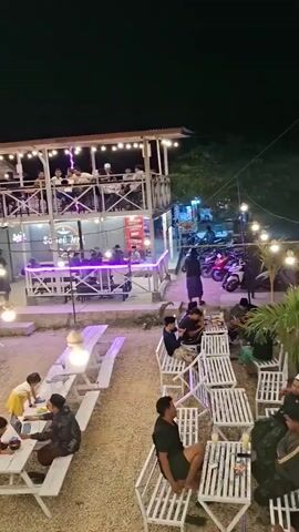 The video shows an outdoor dining area at night. There are multiple levels of seating and dining areas. In the foreground, there are white picnic tables and benches scattered on a gravel surface. Several people are seated at these tables. To the left, a family with two young children is seated at a picnic table. In the middle ground, there is a two-story structure with a white railing and an outdoor staircase. String lights are illuminating the area. People are visible on both levels of the structure, some seated and some standing. To the right, there are several motorcycles parked. Further in the background, there are several small thatched-roof gazebos, also with seating inside, and more string lights. The overall atmosphere appears to be casual and social, with people dining and conversing. The lighting suggests it is nighttime or evening.