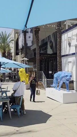 A man in a black t-shirt and blue jeans walks towards the camera, holding a pizza slice in his right hand and a shopping bag in his left. He is eating the pizza as he walks. He passes a blue geometric sculpture of a jaguar, and behind it, a yellow sculpture of a tiger. In the background, palm trees and a building with "SILVERLAKE RAMEN" written on it are visible. To the left, a person sits at an outdoor table under a blue umbrella, with blue chairs and tables. The scene is outdoors on a sunny day.