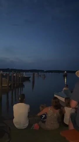 The video begins with a wide shot of a calm body of water at dusk. In the foreground, a group of people, seen from behind, are sitting on the grassy shore, facing the water. A man in a white t-shirt and a baseball cap sits on the left. To his right, a woman in a patterned dress is holding a young child, and next to her is another child. A portion of a person in a hat is visible on the far right. Several boats are docked on the left side of the frame, with posts extending into the water. In the middle ground, several small boats are scattered on the water, some with people in them. In the background, a tree-lined shore is visible, with distant lights suggesting a populated area. The sky is a deep blue with some clouds. The video then zooms in slightly on the water and the boats, with the people in the foreground remaining mostly stationary. The lighting suggests it is early evening.