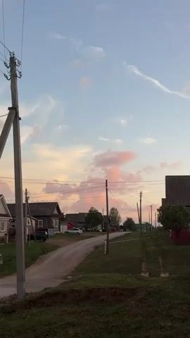 The video captures a rural village scene at dusk. The sky is a pale blue with wispy clouds tinged pink by the setting sun. A dirt road winds through the village, lined with traditional wooden houses and a few scattered trees. Utility poles with overhead wires are visible along the road. A few cars are parked or driving on the road. The overall atmosphere is peaceful and serene.