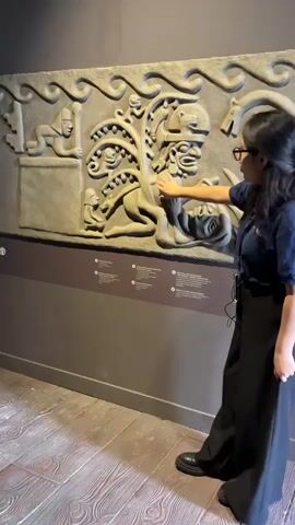 In an indoor setting, likely a museum or gallery in Pueblo Libre, Peru, a woman is seen standing next to a large stone relief artwork. She is wearing a dark blue polo shirt with a visible "LARCO" logo, black wide-leg pants, and black shoes. She has glasses and a lanyard with an ID badge and a small electronic device.

The woman is actively gesturing with her right hand towards the intricate gray stone relief, which features various figures and decorative elements. She appears to be giving a presentation or guiding a tour, speaking enthusiastically and smiling. Her left hand is resting by her side.

The relief is mounted on a dark gray wall. Below the artwork, several small white informational plaques with text and icons are visible, though their content is unreadable. The floor is made of light brown, textured wood or a material resembling wood. The lighting is bright, illuminating the scene and the artwork clearly. The overall atmosphere suggests an informative and engaging cultural experience.