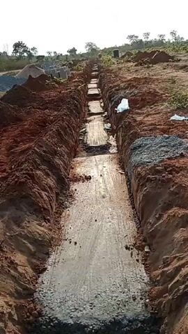 The video shows a long, narrow, partially filled trench in a reddish-brown soil landscape. The trench appears to be for the foundation of a building. Piles of soil flank either side of the trench, with a layer of gray material partially covering the base of the trench. In the distance, there are stacks of rectangular gray blocks. The landscape is generally flat with scattered vegetation. The sky is a hazy white. The video is accompanied by audio stating that the construction is for an airport in Kogi State, Lokoja.
