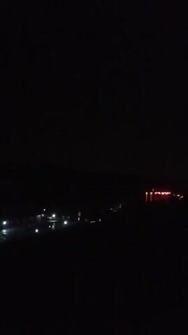 A low-light video shows a rural landscape at night. In the distance, a cluster of buildings is illuminated by bright lights, suggesting a settlement or industrial area. To the right, a larger structure glows with an orange light, possibly indicating activity or decorative lighting. The foreground is dark, obscuring details of the terrain. The overall scene depicts a sparsely populated area at night with pockets of artificial illumination.