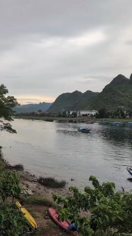 A wide shot shows a river with a sandy bank in the foreground. On the bank, there are two kayaks, one yellow and one red. In the river, there are several boats, some of which appear to be small fishing boats. In the distance, there are green, tree-covered mountains under a cloudy sky. Buildings are visible along the far bank of the river. The lighting suggests it is either dawn or dusk.