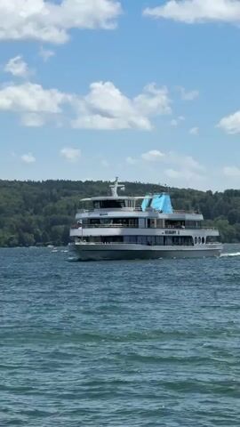 A white passenger boat with multiple decks is sailing across a blue body of water under a cloudy sky. The boat has many windows, a light gray hull, and a prominent light blue section on its upper deck. In the background, a lush green, tree-covered hillside is visible. The water has moderate waves. The camera is positioned low, capturing the boat from a side-on perspective as it moves to the left.