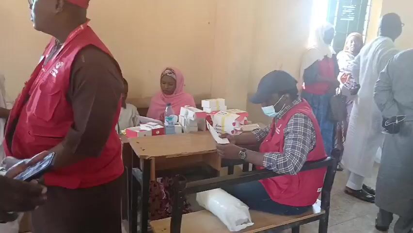 The video shows an indoor scene, likely a clinic or registration area, with several people.  In the foreground on the left, a man wearing a red cap and a red vest with the "Save the Children" logo is looking down at his phone.  To his right, at a wooden desk, a person wearing a black cap, a red vest, and a blue face mask is seated and looking at a document they are holding.  Behind the seated person, stacked on the desk, are several boxes, some white with red markings, and bottles of water.  To the left of the desk, a woman wearing a pink headscarf is also seated.  Further back, several other people are standing and milling about, some wearing headscarves. The walls of the room are a light color. A window with sunlight streaming through is visible on the right side of the frame.  A white plastic bag is on the floor near the seated person at the desk.
