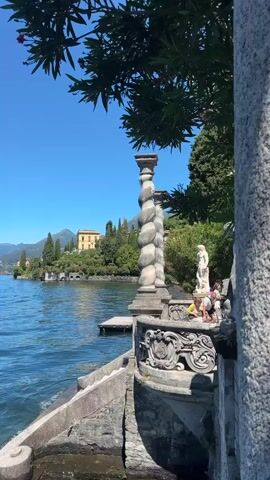 The video opens with a close-up of a stone balustrade on the right, with an ornate pillar in the foreground. The balustrade overlooks a large body of water, likely a lake, with gentle ripples on its surface. Beyond the water, on the opposite shore, is a lush green landscape dotted with trees and a prominent yellow building. The sky is a clear, bright blue. As the video progresses, the camera pans slightly to the left, revealing more of the lake and the distant shoreline. A wooden pier extends into the water from the left side of the frame. Two people are visible in the middle ground, standing near the balustrade. The camera then continues to pan left, capturing a wider view of the lake, the opposite shore with its buildings and trees, and the distant mountains under the vast blue sky. The shot ends by showing the lake stretching towards the horizon.