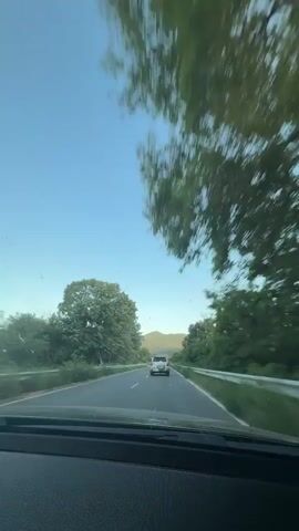 The camera is inside a car, looking out the windshield at a car in front of it on a road. There are trees on either side of the road and mountains in the distance. The sky is blue and clear. The car is traveling on a two-lane road.