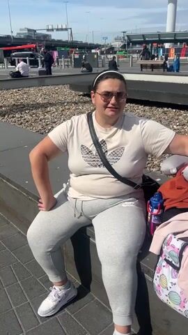 A woman sits on a concrete ledge outside of an airport or bus terminal. She wears a white t-shirt with an Adidas logo, grey sweatpants with a Nike logo, white sneakers, and sunglasses. She has a black purse hanging from her shoulder. To her right is a water bottle and a backpack. In the background are buildings, buses, and other people. She is speaking, likely to someone off-camera.