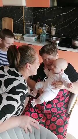 This media file captures a heartwarming family moment in a kitchen setting in Trubacheyevka, Russia. The primary subjects are three individuals: a grandmother holding a baby, a younger woman interacting with the baby, and a young boy standing behind them.

The grandmother, with short, curly hair, is seated and cradling a baby dressed in a white onesie. The baby, with a round face and alert expression, is looking towards the younger woman. The younger woman, with her hair tied back, is leaning in towards the baby, smiling and appearing engaged in a playful interaction. Her attire features a black and white patterned top. The young boy, wearing a striped t-shirt and shorts, stands slightly behind the group, observing the scene.

The background reveals a kitchen environment with peach-colored cabinets and a black marble-patterned backsplash. Various kitchen items are visible, including bottles, containers, and pots on a stovetop, suggesting a domestic, lived-in space. The lighting indicates it's daytime.

The activity captured is a moment of familial affection and connection. The smiles and body language of the younger woman and grandmother convey warmth and joy. The interaction between the woman and the baby, as well as the boy's presence, emphasizes the family bond. The baby's gaze and outstretched hands suggest curiosity and engagement with the people around them.

There is no visible text in the media file. The overall impression is one of a happy, casual family gathering.