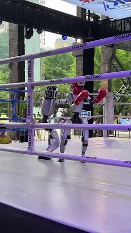 Two humanoid robots are engaged in a boxing match inside a boxing ring. The robot on the left is primarily silver and white with black accents and is wearing black boxing gloves. The robot on the right is also silver and white, with red boxing gloves and a red headpiece. The ring has white ropes and a white canvas floor. The background shows an outdoor setting with trees, buildings, and a crowd of people watching the event. There are banners and lighting rigs above the ring. The robots are moving and appear to be punching each other.