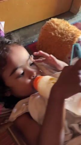 A child is lying down and drinking from a baby bottle while looking at a smartphone. The child is wrapped in a blanket and appears to be resting on a pillow. The background is blurred but suggests an indoor environment with a door and a rug.