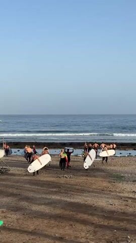 This media file depicts a group of people, likely a surf school, on a sandy beach in Arona, Spain. The scene is set at the shore of the ocean, with gentle waves rolling in under a clear, pale blue sky, suggesting it is morning or late afternoon. The weather appears to be warm and pleasant.

The primary subjects are approximately two dozen individuals, mostly men, some of whom are wearing wetsuits and carrying surfboards. They are scattered across the beach, with some walking towards the water and others gathered in small groups. Several people are carrying white surfboards horizontally above their heads or vertically at their sides, indicating they are preparing for or have just finished a surfing session. One person in a black wetsuit is walking away from the camera towards the left side of the frame, while others are facing the ocean.

The objects visible include numerous surfboards of varying sizes and colors, some of which are white, and a few are dark. There is also a metal structure, possibly part of a surf school setup or a beach equipment rack, on the far left of the image.

The overall activity appears to be a surfing lesson or a group surfing excursion. The atmosphere seems relaxed and active, with people engaged in preparing for or participating in water sports. No discernible text is visible in the media file. The location is suggested to be a beach in Arona, Spain, known for its coastal activities.