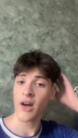 A young person with dark, styled hair is looking into the camera and touching their hair. They are wearing a blue shirt. The background appears to be a wall with a mottled green pattern.