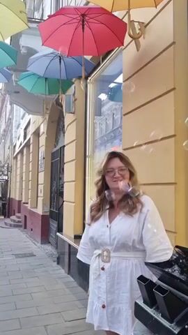 A woman wearing glasses and a white dress stands on a cobblestone sidewalk. She is smiling at the camera. Behind her, colorful umbrellas are suspended overhead. Bubbles are floating in the air around her. The scene appears to be an outdoor street or marketplace.