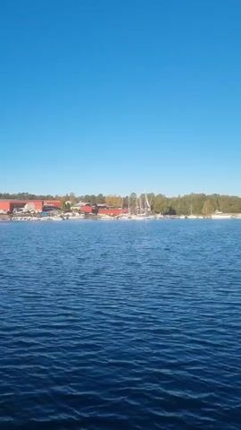 The video shows a body of water, likely a lake or bay, under a clear blue sky. The water is rippled with small waves. In the distance, there is land with buildings, including one that is large and red, as well as trees and some boats docked along the shore. As the camera pans slightly, the view expands to show more of the waterway and the opposite shoreline with denser foliage.