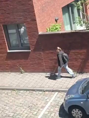 A person walks on a paved sidewalk next to a red brick building. The person is wearing a dark jacket, light-colored pants, and carrying a backpack. The person walks towards the building and then appears to turn and walk away from the camera. The scene is outdoors, with a tree and some plants visible.