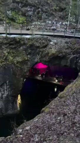 The video shows a rocky quarry with a cave containing a man-made deck inside, where people are gathered. The cave is illuminated by colored lights, with the colors changing between red, blue, and green. Water is visible in the quarry below the cave. A wooden walkway with railings is above the cave on the left. The environment is rocky with some moss and vegetation. The video appears to have been taken outdoors in daylight.