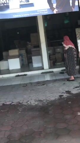 The video shows a woman in a pink headscarf and a patterned dress standing outside a store. The store appears to sell tiles, with stacks of boxes visible inside. The ground outside the store is paved with hexagonal tiles and is wet, possibly from rain. The woman is looking into the store. The lighting suggests it is daytime.