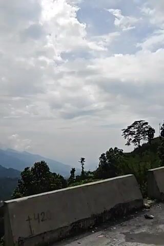 The video shows a view of a mountainous landscape with clouds. In the foreground, there are concrete barriers and lush green foliage, including trees. The camera appears to be moving, possibly from a vehicle, as the perspective shifts across the scene. The mountains in the background are covered in trees and shrouded in mist or clouds. The sky is overcast with dramatic cloud formations.