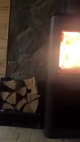 A static medium shot shows a modern, black wood-burning stove, with a clear glass door, burning brightly inside. A stack of split firewood sits to the left in a black metal rack. The stove stands against a wall partially covered with rough, gray stonework. The floor is light gray and has the appearance of wood-look tile. The environment is dimly lit.