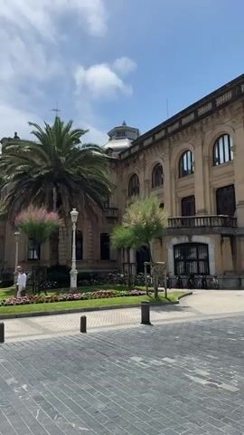 A man walks across a paved area in front of a tan building with arched windows and a palm tree. Another person is visible on the right side of the building. A motorcycle passes by in the middle ground. The sky is blue with white clouds.