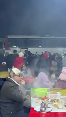 The video shows a bustling outdoor area with people dining at tables, surrounded by smoke or mist. Several individuals are seated, enjoying food and drinks at red and yellow tables. Behind them, a crowd gathers near a white bus, with some people moving around. A man in a green bucket hat is prominent in the foreground. The atmosphere appears lively and possibly festive, with background music adding to the scene. Towards the end of the video, a man in a tan coat is seen walking towards an area where carcasses are hanging, suggesting a cooking or grilling setup.