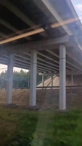 The video captures a view from a moving vehicle, likely a train or car, looking outwards. The camera pans across a landscape featuring a concrete overpass with thick supporting pillars visible in the foreground. Beyond the overpass, a ramp leads up to the elevated road, lined with trees. The sky is partly cloudy, and sunlight intermittently brightens the scene, creating lens flare in some shots. The surrounding area is green with grass and various types of trees, suggesting a rural or semi-rural environment. There are no people or animals visible in the video. The action is limited to the continuous movement of the vehicle and the changing perspective of the scenery.