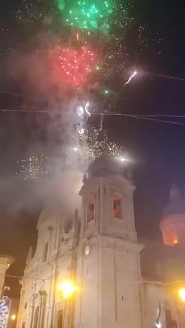 This is a video of fireworks exploding in the night sky above a white building that appears to be a church or cathedral, with a tall steeple and dome. The fireworks are various colors, including white, red, and green, and they are illuminating the sky and the building below. The video is shaky, suggesting it was filmed by someone holding a phone. The footage is taken at night.