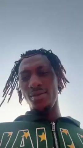 The video shows a person with dreadlocks wearing a green hoodie. They are looking up at the sky. The sky is a light blue color, possibly indicating an overcast day or early morning. The person looks back down at the camera and smiles slightly.