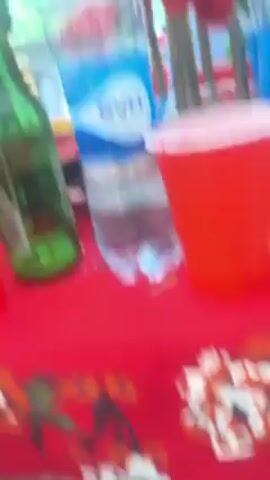 The video shows a table at an event with a red tablecloth. On the table are two red plastic cups, a green beer bottle, and a bottle of "eva" water. A man in a red hat and sunglasses is visible in the background, pointing. Other people are visible in the background as well. There are also red flowers on the table.