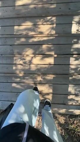 The video shows a first-person perspective of someone walking on a wooden bridge. The person is wearing light-blue jeans and black and white sneakers. A black bag hangs around their body. The bridge has wooden planks as its surface and wooden railings on the sides. Shadows from trees or foliage are cast onto the bridge. Green vegetation can be seen below the bridge. The video is shaky, indicating that it was shot while the person was moving.