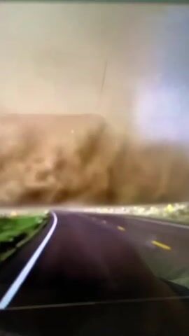 A tornado is visible in the distance, approaching what appears to be a road. The tornado is a funnel cloud made of dirt and debris, indicating a dust storm or a powerful wind event. The road is paved and has lane markings, suggesting it is a highway. The surrounding environment is rural, with green grass and possibly some vegetation visible on the sides of the road. The sky is overcast with a hazy, brown hue, characteristic of a dust storm. The video seems to be captured from a moving vehicle, as the perspective suggests forward motion.
