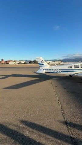 A drone captures an aerial view of two small airplanes parked on an asphalt tarmac under a clear blue sky. One plane, predominantly white with blue accents and the letters "LA" on its tail, is positioned closer to the camera. The second plane, also white, is further away, with its tail visible. A man in a dark t-shirt and pants is seen interacting with a refueling station in the foreground, with the sun creating a lens flare as it shines from behind him. The environment appears to be an airfield, with buildings and distant hills visible in the background. Shadows of people and possibly the drone are cast across the tarmac.