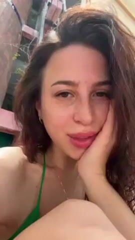 A young woman is shown in a close-up selfie shot. She has light skin, brown hair, and is wearing a green swimsuit. She is smiling slightly and resting her cheek on her hand. A building or structure is visible in the background. The lighting is bright, suggesting an outdoor setting.
