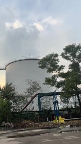 The video shows a series of large, white industrial storage tanks against a cloudy sky. The tanks appear to be connected by elevated walkways. Dense green foliage and trees surround the base of the tanks, with some debris visible in the foreground. A section of piping, painted red and blue, is also visible attached to one of the tanks. The lighting suggests it is either dawn or dusk, with the sun peeking through the clouds. The camera pans slowly across the scene.