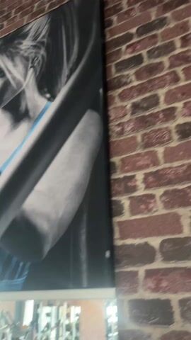 A close-up shot, taken from a low angle, pans across a mural in what appears to be a gym. The mural features black and white images of muscular figures, interspersed with a single color element - a bright blue sports bra worn by one of the female figures. The gym environment is subtly visible through a large mirror running below the mural, showcasing various exercise machines. The mural is mounted against a brick wall with an uneven surface. The primary focus is on the juxtaposition of the monochrome figures and the vibrant blue color accent.