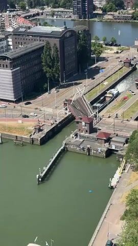 This video captures an aerial view of a bascule bridge in operation, likely in an urban setting with a canal or waterway. The bridge is partially raised, allowing a boat to pass underneath. A small control tower is visible next to the bridge, with people standing nearby. There is traffic on the road adjacent to the bridge, including cars and trams. Trees and buildings are visible in the background, suggesting a populated area. The water is a greenish hue, and the sky is clear and blue. The overall scene depicts a functional piece of urban infrastructure in use.