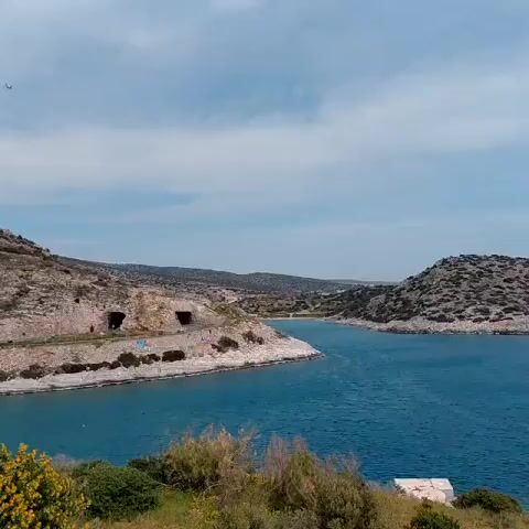 The camera pans across a coastal scene, featuring a body of water, rocky peninsulas, and a partly cloudy sky. On the left peninsula, there are openings resembling tunnels or caves, with some graffiti visible along the lower edge. The water appears blue and calm. The right side of the view reveals a rocky hill and a glimpse of a distant town or settlement nestled along the coast. In the foreground, there's green vegetation and a small, white rectangular structure or object on the grassy land.