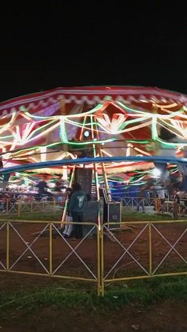 At night, a carnival carousel is in motion, adorned with bright, colorful lights. The carousel’s top is a red and white striped tent. People stand around the ride, some watching and others appearing to be part of the setup or operation. The carousel is surrounded by a yellow metal fence. The ground inside the fence is dirt and some patches of green grass. The overall environment suggests an outdoor fair or carnival at night.