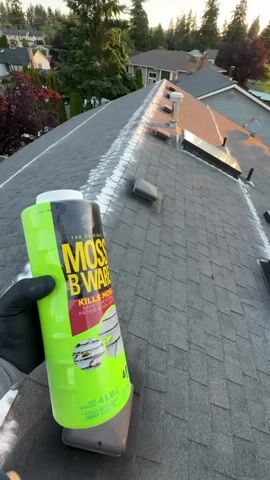 The video shows a close-up of a person's gloved hand holding a can of "MOSS B GONE" product on a rooftop. The can is green and yellow with red lettering. The rooftop is covered in asphalt shingles and has several skylights and vents. White granules are visible on the roof, particularly around the skylights, indicating the product has been applied. The camera pans across the rooftop, revealing a suburban neighborhood with houses and trees in the background. The sun is setting, casting a warm glow on the scene.
