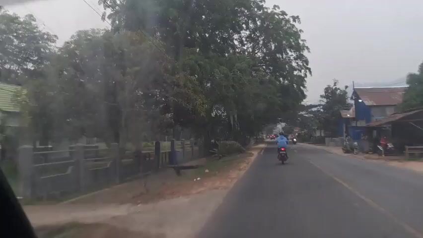 A dashcam view captures a view from inside a vehicle, looking out through the windshield. The vehicle is traveling on a paved road, which is wet, suggesting recent rain or a damp environment. The road is bordered on the left by a dirt shoulder and a fence with trees and foliage beyond it. On the right, the road is lined with buildings and more trees. Several motorcycles are visible on the road, with one in the foreground on the right and another further ahead on the left. The sky appears overcast, with a light grey hue. The overall scene suggests a rural or semi-urban environment during daylight hours, possibly early morning or late afternoon due to the soft lighting.