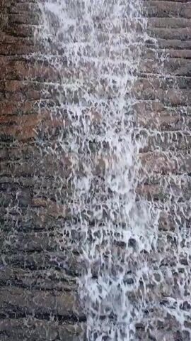 The video displays a close-up view of water cascading down a terraced or stepped wall. The water is white and frothy, indicating a continuous and active flow as it tumbles over multiple horizontal layers. The structure beneath the water appears to be made of brick-like or stone-like material, possibly concrete, arranged in a stepped pattern. No people or visible text are present. The scene suggests an artificial waterfall or water feature, likely part of a landscape design in an urban or park setting. The lighting indicates it is daytime.