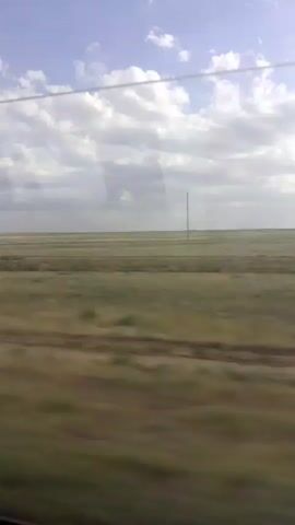 The video shows a view from inside a vehicle, likely a train, looking out a window at a vast, flat landscape. The landscape appears to be a prairie or grassland, with low vegetation in shades of green and brown. The horizon is clearly visible, meeting a sky filled with scattered white clouds against a pale blue background. In the distance, a single, tall, thin pole stands vertically against the sky. The movement of the vehicle is evident in the motion blur of the foreground and midground, indicating travel. No people or distinct objects other than the distant pole are discernible. The overall impression is one of open space and movement through a rural environment.