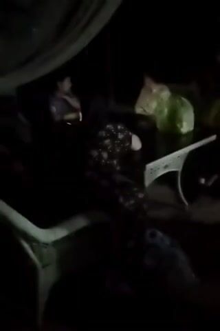 The video appears to be filmed at night or in a dark environment. Several people are visible, seated around a table. One person, a woman in a floral dress, is leaning back in her chair and looking to her right, with her hand touching her head. Another person, wearing a blue shirt, is looking down at a device in their hands. There are also other individuals partially visible in the background. Objects on the table include what appear to be plastic bags, one of which is green. The lighting is dim, suggesting it could be from a phone or a small light source. The overall atmosphere seems casual, possibly a social gathering.