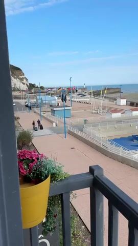 The video shows a boardwalk or promenade overlooking a beach and the sea under a clear blue sky with some clouds. In the foreground, there is a railing with a yellow pot containing pink and green flowers. To the left of the promenade, there are flags on poles, and a white lighthouse can be seen in the distance. Further down the promenade, there are beach huts and what appears to be a children's play area with blue surfaces and climbing equipment. Some people are visible walking on the promenade, and a few are in or near the water on the beach. There is also a concrete pool or water feature with people in it. In the middle ground, there are several small white structures with red roofs, possibly changing rooms or storage sheds. The sea appears to be at low tide, with a wide expanse of wet sand visible. A few people are scattered on the beach. The overall scene is one of a seaside resort on a sunny day.