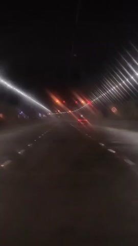 This is a first-person perspective video, likely from a vehicle, traveling through a tunnel at night. The tunnel has bright white lights along the left side and a series of orange lights, some with red X's, along the ceiling in the distance. The road has white dashed lines and reflective markers. There is some atmospheric haze or mist, causing light streaks.