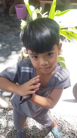 A young boy with black hair and a grey t-shirt and shorts is standing on a gravel surface outdoors. He is holding a small object, possibly a phone, in his hands and looking at it. There are plants and a concrete planter in the background. Sunlight is casting shadows on the ground.