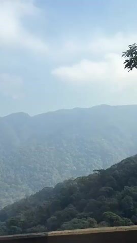 The video shows a view from a moving vehicle overlooking a lush, green, mountainous landscape under a hazy sky. The foreground is dominated by a concrete barrier, suggesting the viewpoint is from a road or highway. The mountains are densely covered with trees, creating a textured, undulating surface. The sky is light blue with scattered, wispy clouds. The overall impression is one of natural beauty and a sense of travel.