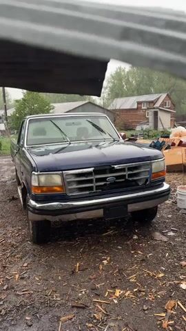 The video shows a navy blue Ford F-series pickup truck parked on a muddy ground in a rural setting. It is raining. The truck is facing the camera, and the windshield wipers are raised. In the background, there is a wooden house with a metal roof, along with various other buildings and trees. The ground around the truck is covered in wood chips and debris, and there are cardboard boxes, a bucket, and other objects scattered nearby. The video is shot from under a metal roof, providing a partial frame at the top.